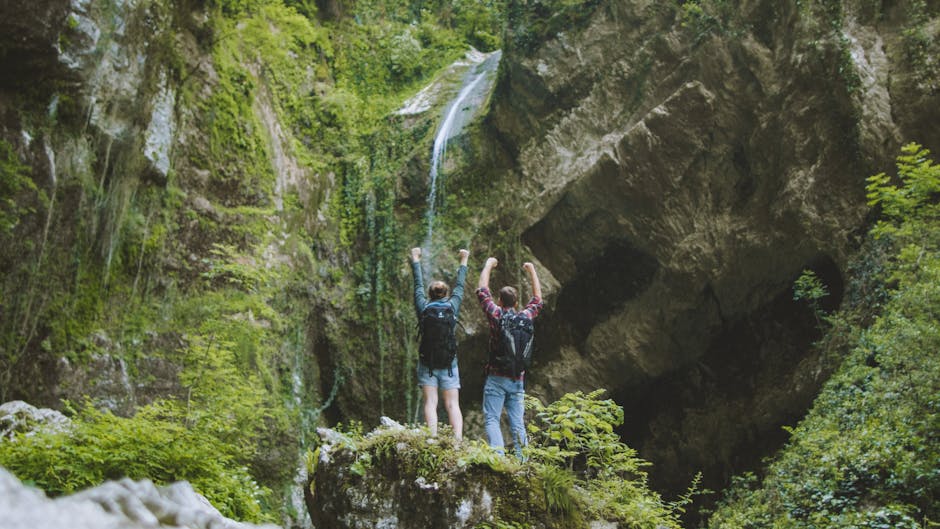 A joyful couple with backpacks raising arms in victory in front of a waterfall amidst lush greenery
